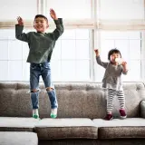 Dos niños saltan en un sofá. El mayor, mi hijo de 2 años, ríe con los brazos en alto. La pequeña bebe de un biberón.
