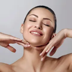 Rostro de mujer con ojos cerrados y sonrisa suave, manos acariciando su cuello. Piel radiante y matificante.