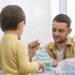 Un niño de 3 años, con tartamudeo, juega con bloques. Su padre lo mira con ternura mientras construyen juntos.
