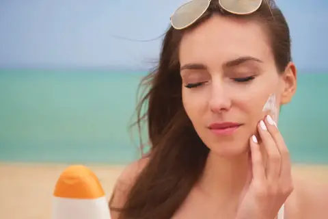 Mujer se aplica protector solar en la cara en la playa. El protector solar hidrata la piel mientras la protege del sol.