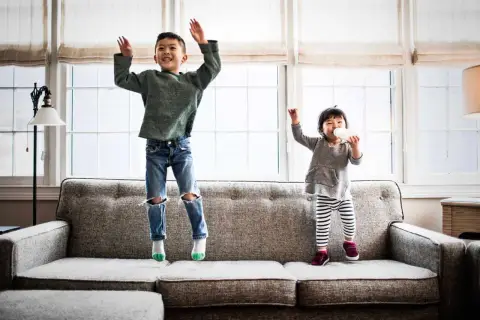 Dos niños saltan en un sofá. El mayor, mi hijo de 2 años, ríe con los brazos en alto. La pequeña bebe de un biberón.
