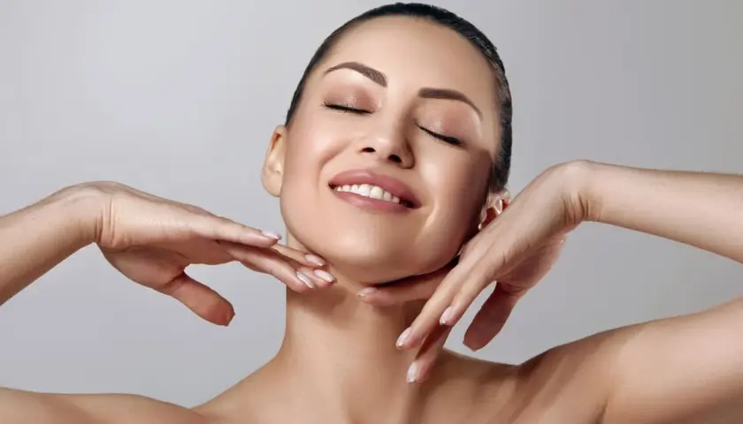 Rostro de mujer con ojos cerrados y sonrisa suave, manos acariciando su cuello. Piel radiante y matificante.