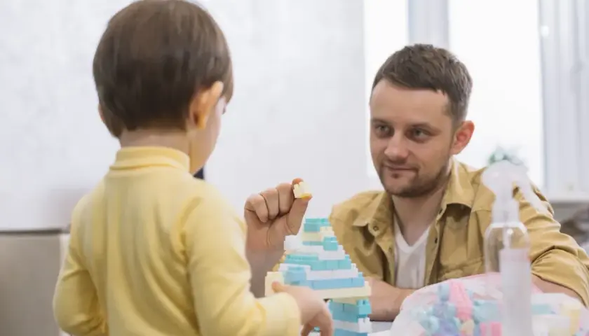 Un niño de 3 años, con tartamudeo, juega con bloques. Su padre lo mira con ternura mientras construyen juntos.