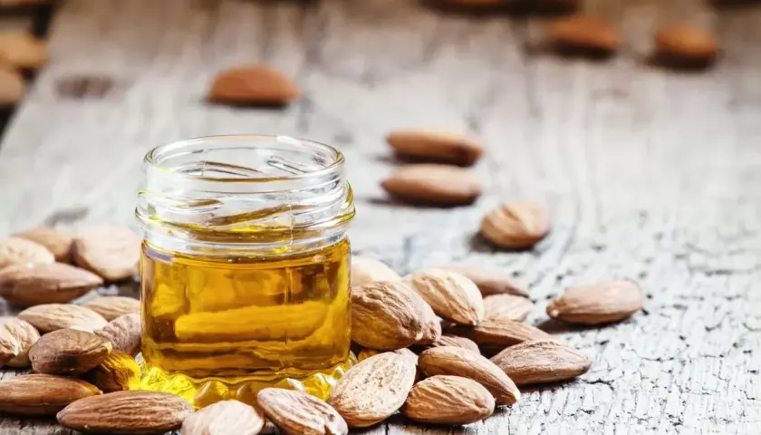 Un frasco de aceite de almendras para la cara rodeado de almendras enteras sobre una superficie de madera rústica.