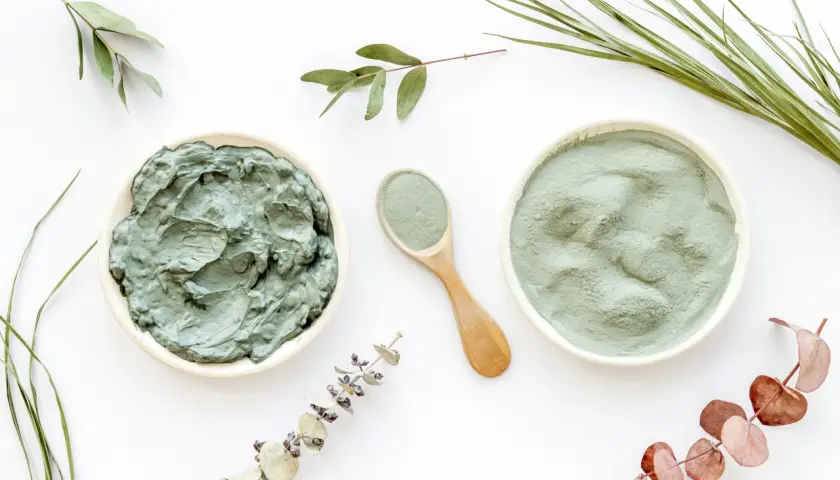 Dos recipientes con arcilla verde, uno en polvo y otro en pasta, junto a una cuchara de madera y ramas secas. Descubre las arcilla verde propiedades.