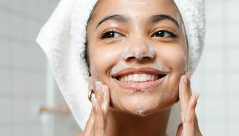 Una mujer sonríe mientras se aplica espuma en el rostro, demostrando en qué consiste la doble limpieza facial para una piel radiante.