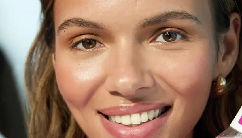Una mujer sonríe, mostrando su piel radiante. Sostiene un producto con agua de rosas, ideal para la doble limpieza facial.