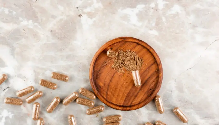 Cápsulas de maca esparcidas sobre un fondo de mármol, con un cuenco de madera que contiene polvo de maca y una cápsula abierta.