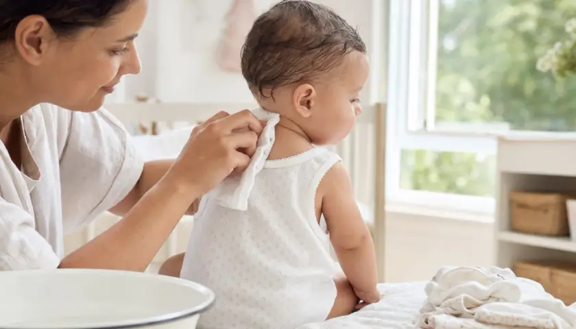 Madre limpia la nuca de su bebé para prevenir la sudamina. El bebé, sentado, mira hacia la ventana.