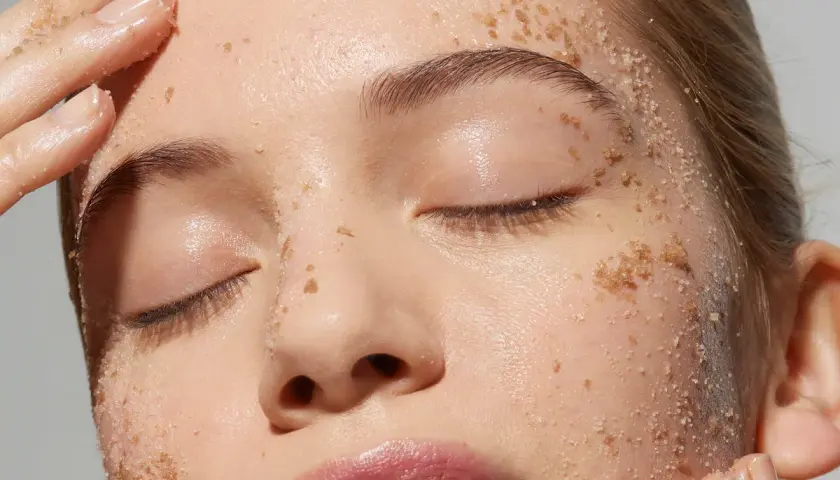 Una cara llena de granos de exfoliante, con dedos aplicándolo suavemente.