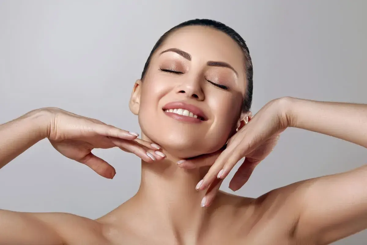Rostro de mujer con ojos cerrados y sonrisa suave, manos acariciando su cuello. Piel radiante y matificante.
