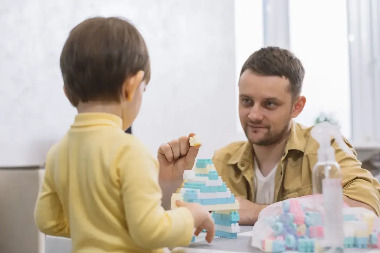 Un niño de 3 años, con tartamudeo, juega con bloques. Su padre lo mira con ternura mientras construyen juntos.