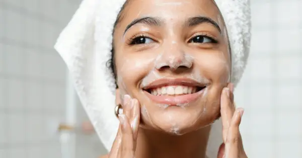 Una mujer sonríe mientras se aplica espuma en el rostro, demostrando en qué consiste la doble limpieza facial para una piel radiante.